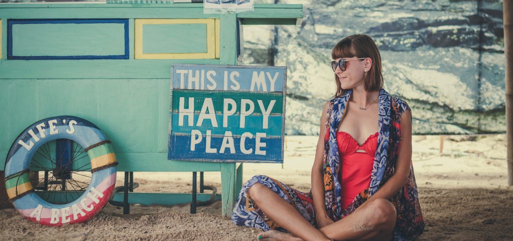 Lady sitting on the beach with a sign that reads "this is my happy place" and "life's a beach"