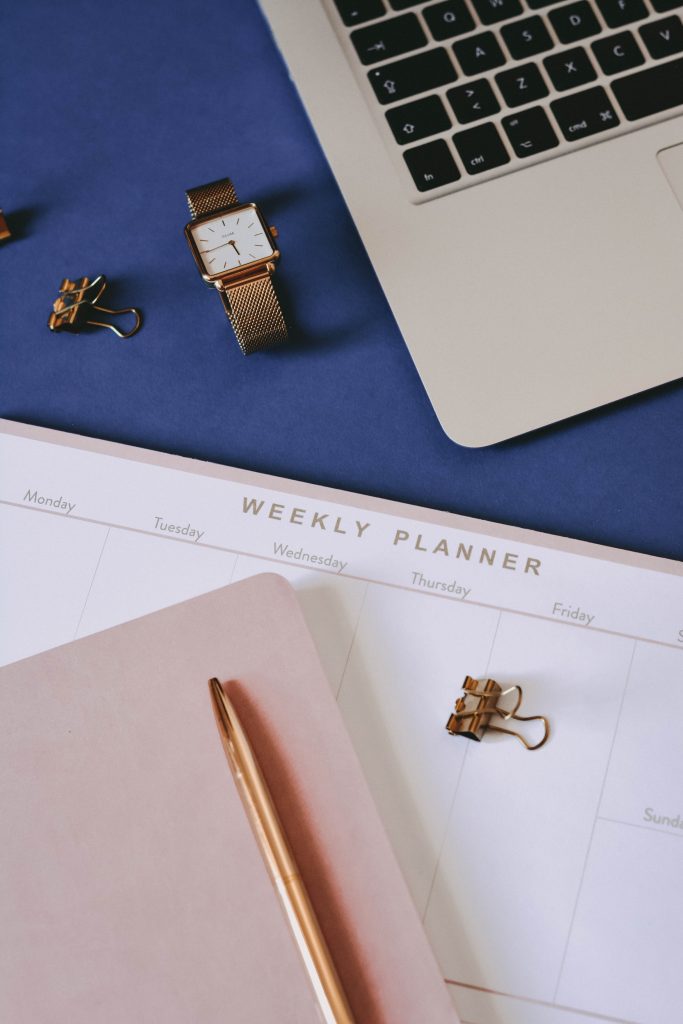 Photo of Weekly Planner with a pink notebook with golden colored pen on top and clip on the right side.