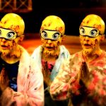 4 yellow faced figures with wearing eyemasks (with pictures of eyes on the masks) stand with their hands together as if praying.