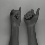 Video still of two forearms. On the hand on the left the little finger is raise and on the one on the right the thumb is protruding will the other fingers are clasped.