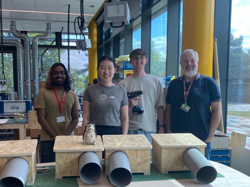 Four people standing and smiling in a workshop with four hedgehog houses in front of them.