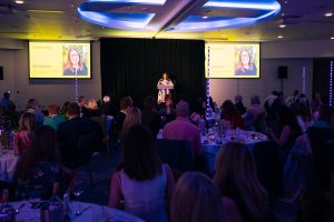 Bec Bennett presenting the Sustainability Champion Award on stage to a seated audience, with her image shown on two large screens in front of a seated audience in a blue-lit room.