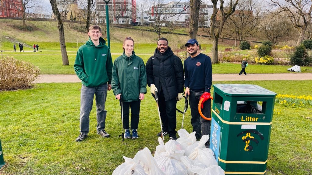 Four volunteers smiling and holding litter pickers, stood besides some bags of rubbish and a bin in Peel Park.