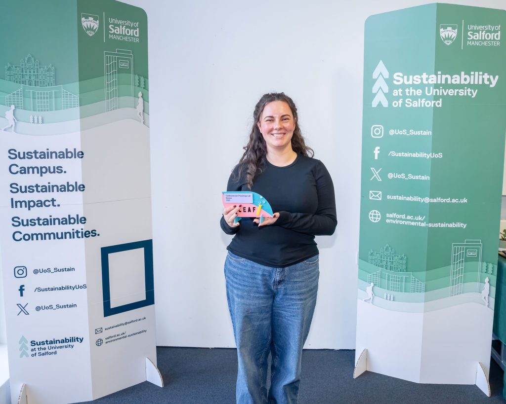 A person stands between two green sustainability banners and smiles as the hold a pink, yellow and blue award.