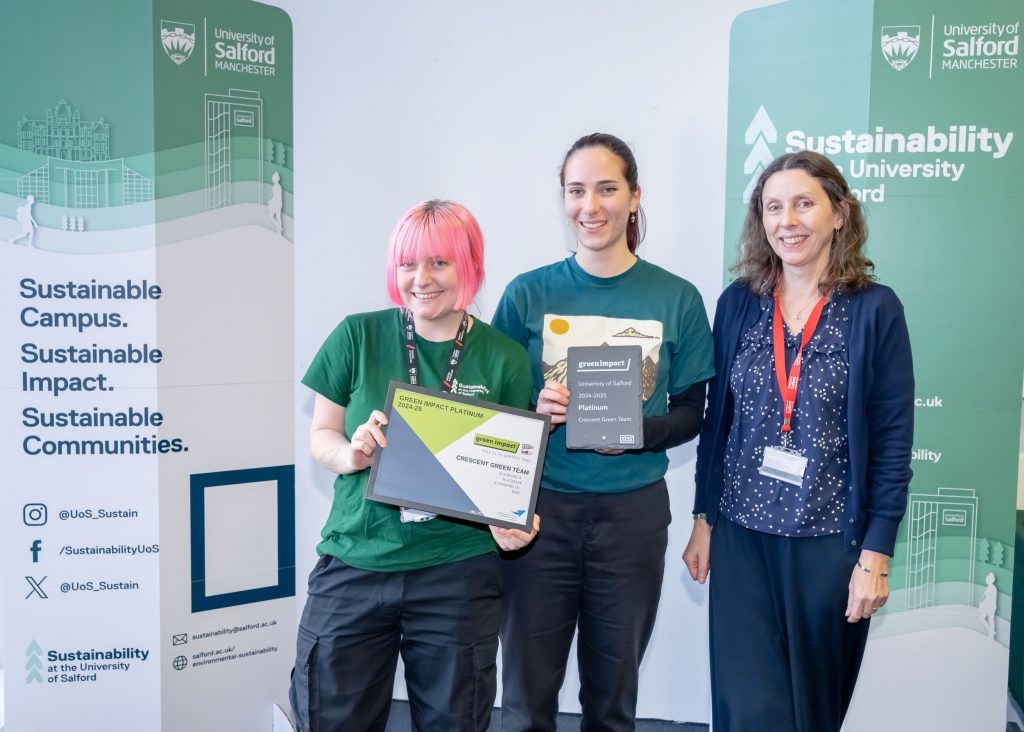 Three people stand smiling in front of two green banners. The person in the middle holds a grey slate plaque and the person to the left holds a framed certificate.
