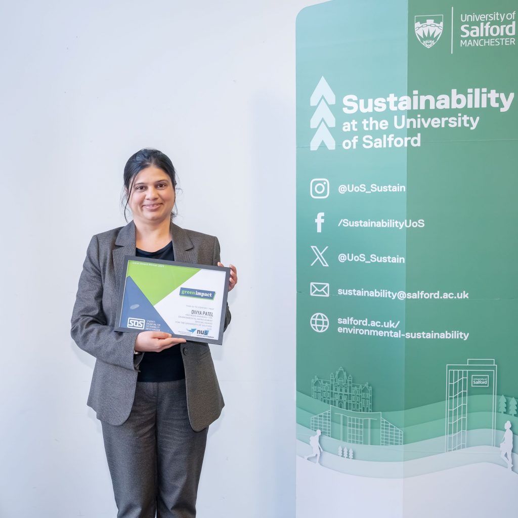 A person stands between two green sustainability banners holding a framed certificate and smiling.