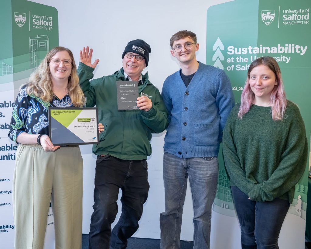 Three people stood in a line smiling. The woman on the far left holds an award certificate and the man to her right holds a slate trophy. They are all stood in front of two green sustainability banners.