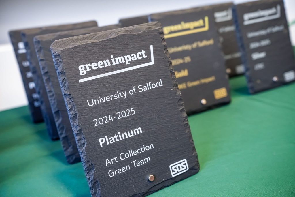 A set of grey slates with the Green Impact award details printed on the front. In the foreground is awards with white lettering saying 'Green Impact, University of Salford, 2024-2025, Platinum. In the background are blurred versions of the award all sat on a green table cloth.