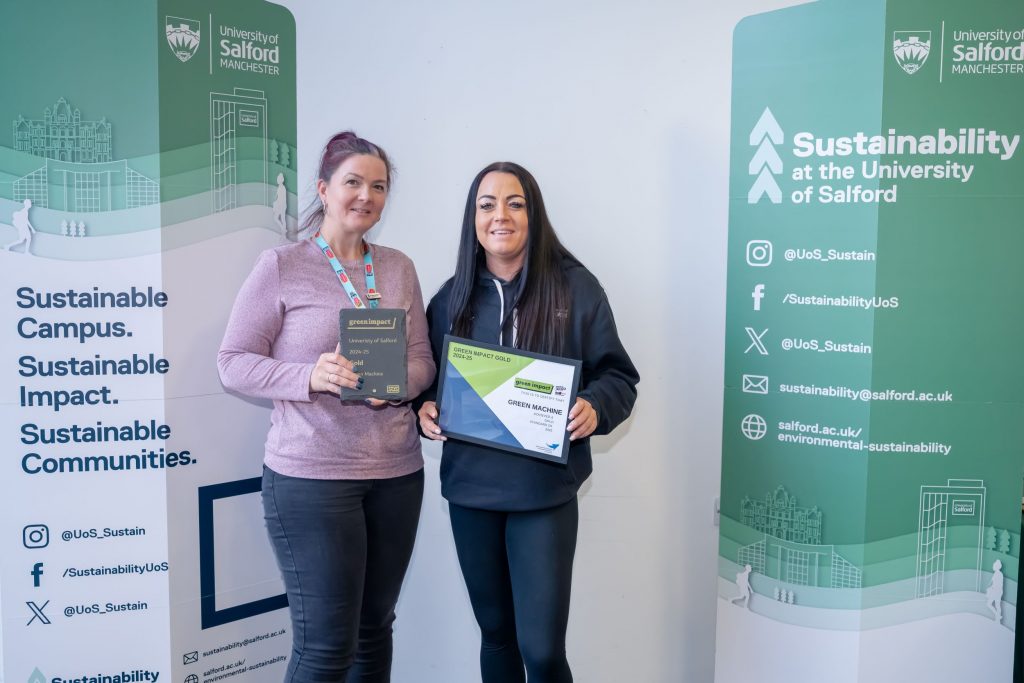 Two people stand side by side in front of two green sustainability banners. The one on the left holds a slate plaque and the one on the right holds a framed award certificate.