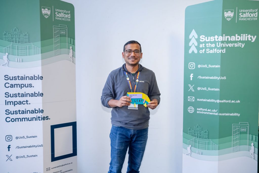 A person stands between two green sustainability banners and smiles as the hold a pink, yellow and blue award.
