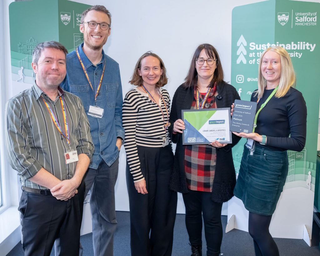 A group of five people stand together smiling in front of two green sustainability banners. The person on the fair right holds a grey slate plaque and the person to their right holds a framed certificate.