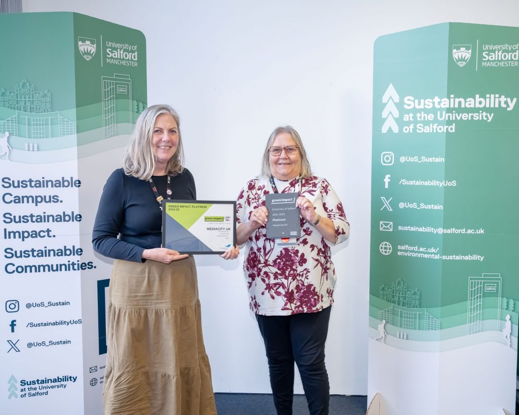 Two people stand between two green sustainability banners. The person on the left holds a framed award and the one on the right holds a grey plaque.