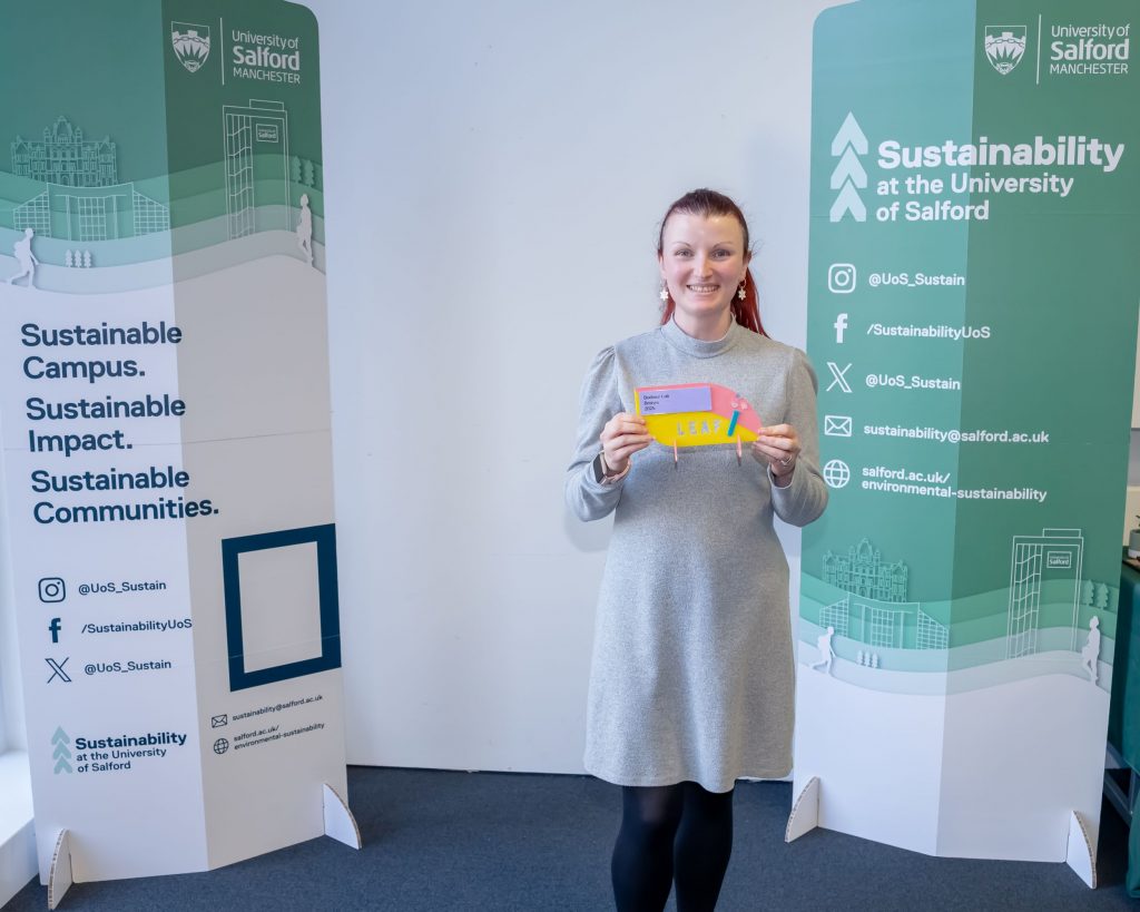 A person stands between two green sustainability banners and smiles as the hold a pink, yellow and blue award.