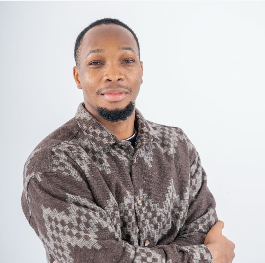A person with short dark hair, a short beard, smiling and crossing their arms, in front of a white background.