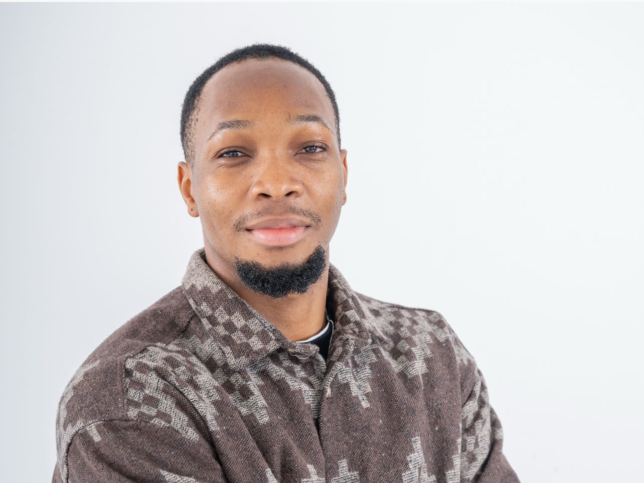 A person with short dark hair, a short beard, smiling and crossing their arms, in front of a white background.