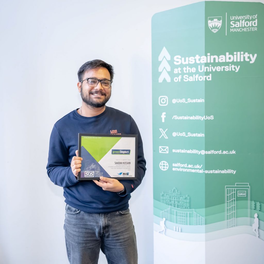 A person stands between two green sustainability banners holding a framed certificate and smiling.