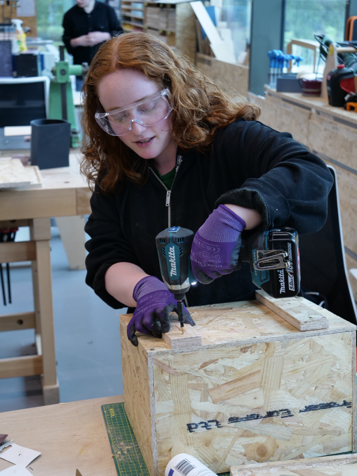 Iona wearing safety goggles and gloves uses a cordless drill to assemble a wooden box in a workshop.