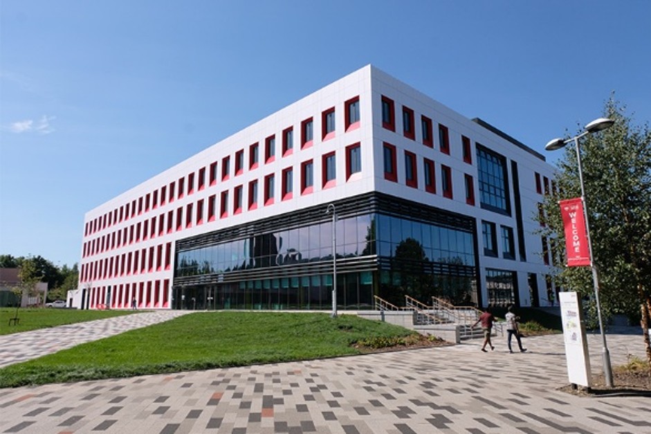 Modern multi-storey academic or office building with a white exterior and red window surrounds. Large glass-fronted ground floor reflects surrounding trees and sky. The building sits beside a landscaped lawn and paved pedestrian walkway, with two people walking near the entrance. A red banner mounted on a lamppost displays the word “Welcome.” Clear blue sky overhead.