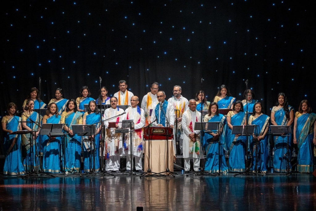 The Indian Choir of England performing in front of a starred black background. Rakesh is at the foreground as Artistic Director. The aim of the image is too show the choir performing and visually demonstrate Rakesh' role in the BVG.