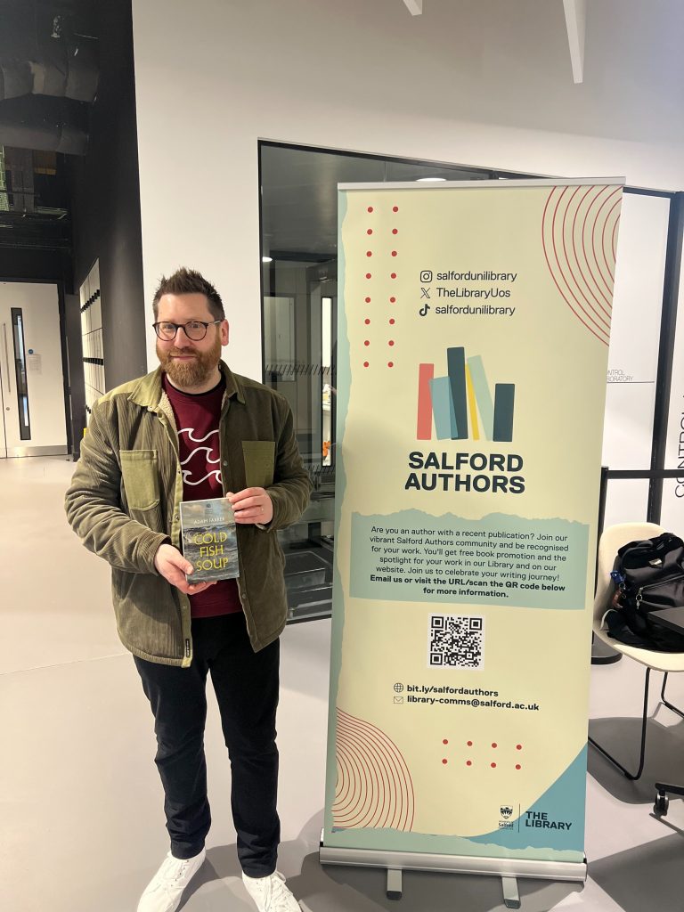 Adam Farrer holding his book infront of Salford Authors Banner