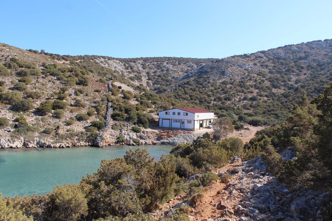A white Greek house with a red roof on a rocky beach next to the ocean with rocky hills behind it