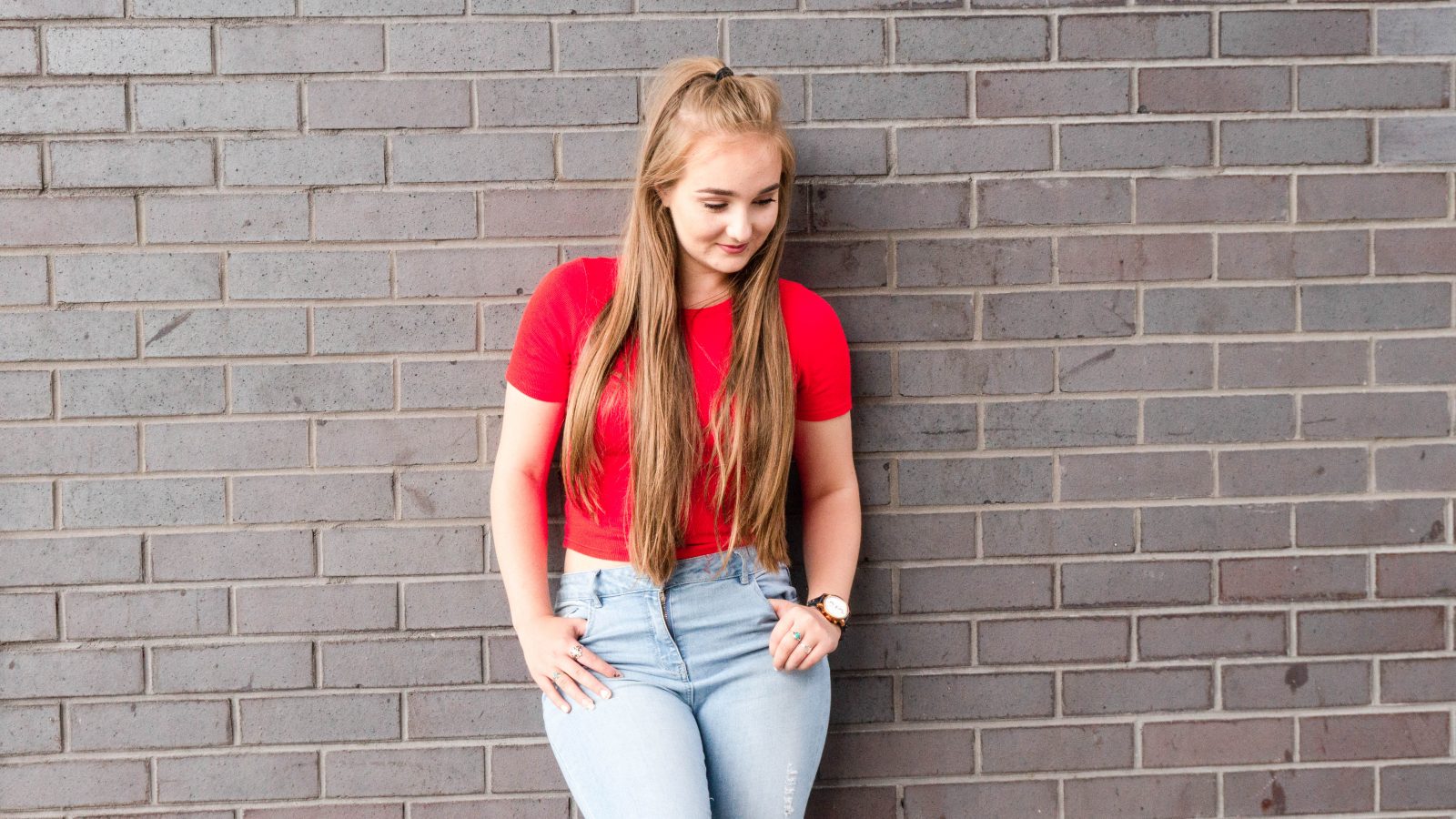 Becky stood next to a dark brick wall with her hands in her pockets looking down