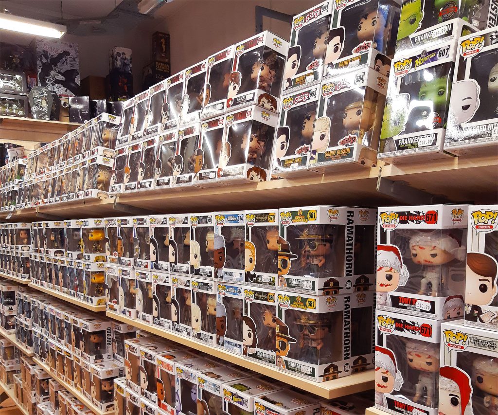 Three rows of wooden shop shelves in the Forbidden Planet Manchester store which are full of Funko Pop Vinyl figures new in their boxes. Nearest to the camera you can see characters from series such as Grease and Die Hard.