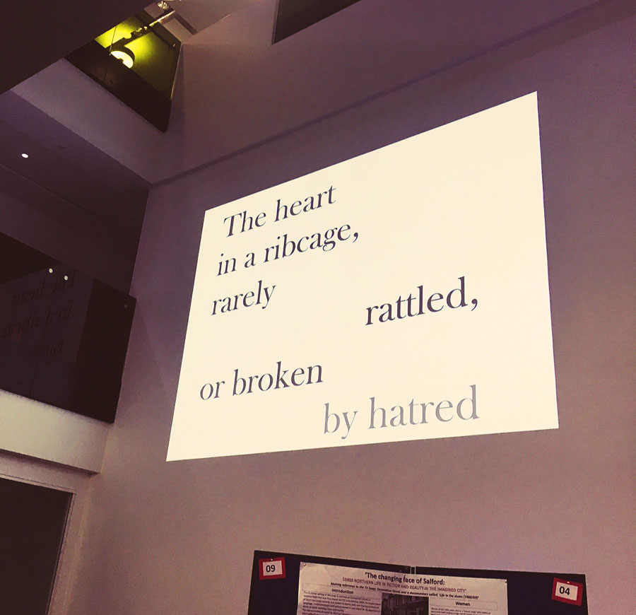 There is a presentation projected onto a blank wall in the University of Salford's MediaCityUK campus. On the projection is some visual poetry spaced out. It reads 'The heart in a ribcage, rarely rattled, or broken by hatred.'