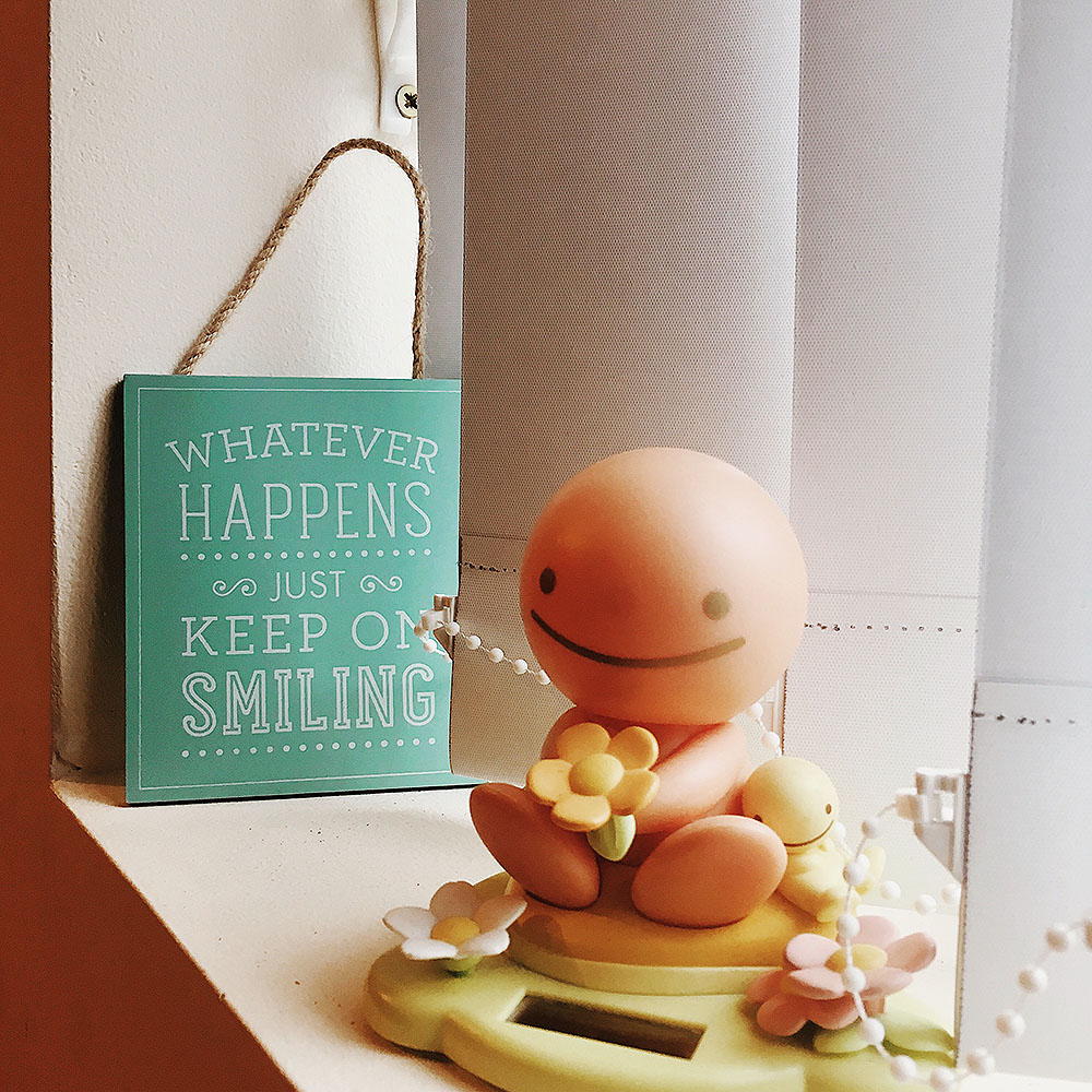 A small wooden hanging that says 'Whatever happens just keep on smiling' next to a solar powered 'Sun Buddy' bobble head. 