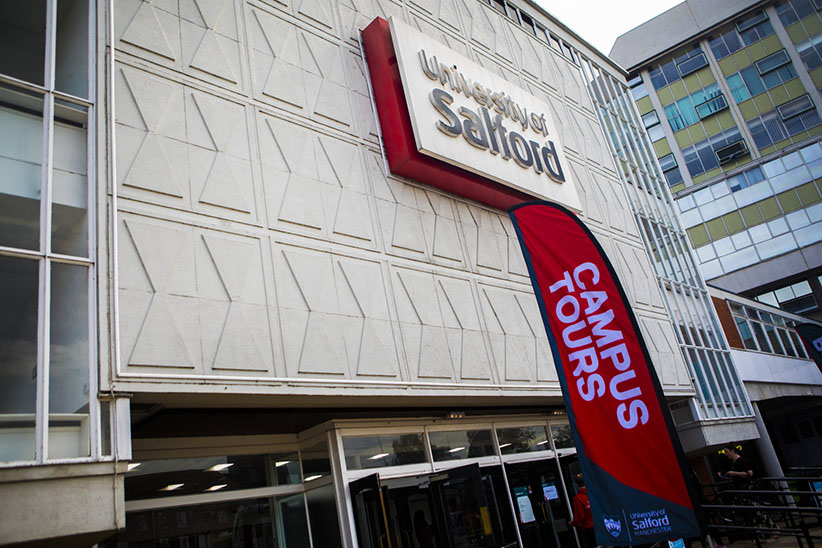 Photo of the front of Maxwell Hall on the main University of Salford campus. You can see our logo on top of the building that reads 'University of Salford'. In the foreground is a temporary sign advertising 'Campus Tours' as this photo was taken at an open day.