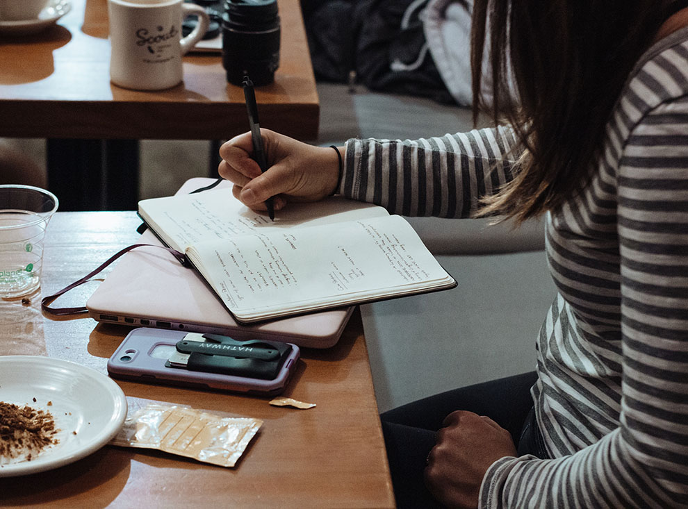 A female in a striped shirt sat at a table in a public space. She has a plate with food crumbs and a drink cup next to her. She is writing in a notebook thats sat on top of her pink laptop.