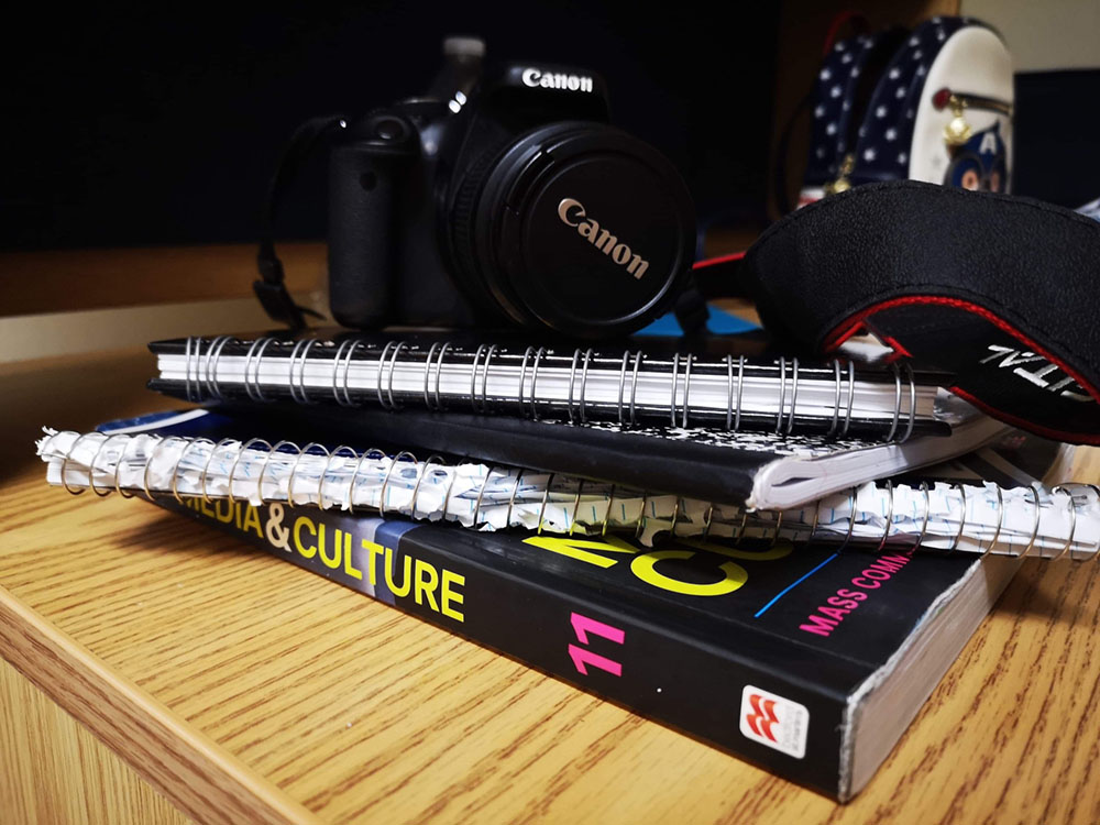 A Canon camera placed on top of several notebooks and spiral bound books. With a backpack in the background.