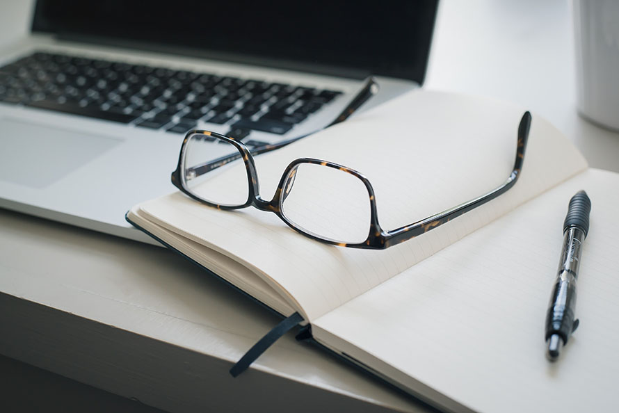 A laptop on a desk alongside an open notebook, glasses and a pen.