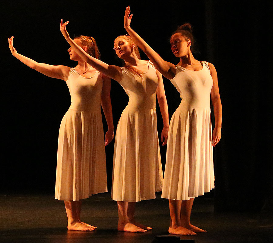 Three of our University of Salford Dance students performing in a warm light on the New Adelphi Theatre stage. In unison the girls are raising their right arms during their dance performance.