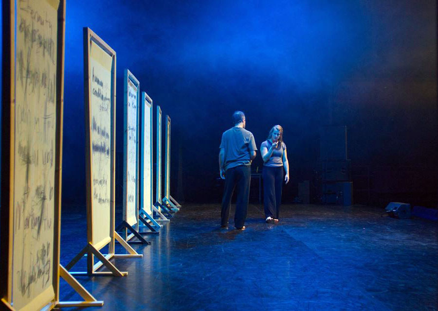 On the left side of the photo we see across a stage is multiple wooden stands that have been written on repeatedly as part of the 'Vital Signs: Wrestling Truth' performance. Sarie Mairs Slee and Scott Thurston are travelling the stage in grey and black clothes.