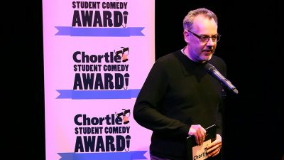 Steve Bennett, manager of chortle hosting on the main stage in front of two flats with the 'Chortle Student Comedy Awards' logos on