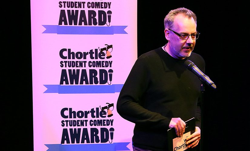 Steve Bennett, manager of chortle hosting on the main stage in front of two flats with the 'Chortle Student Comedy Awards' logos on