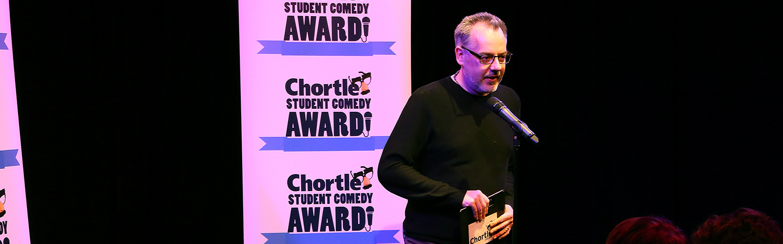Steve Bennett, manager of chortle hosting on the main stage in front of two flats with the 'Chortle Student Comedy Awards' logos on