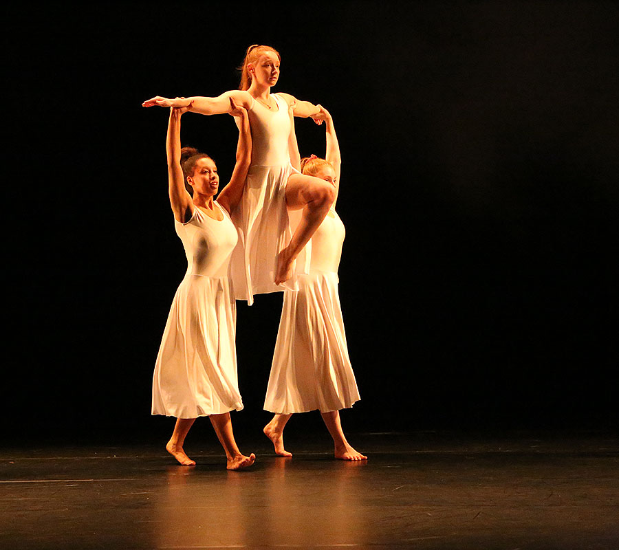 The three Salford dance students performing in a warm light. Two of the girls have the other held up high above their shoulders.