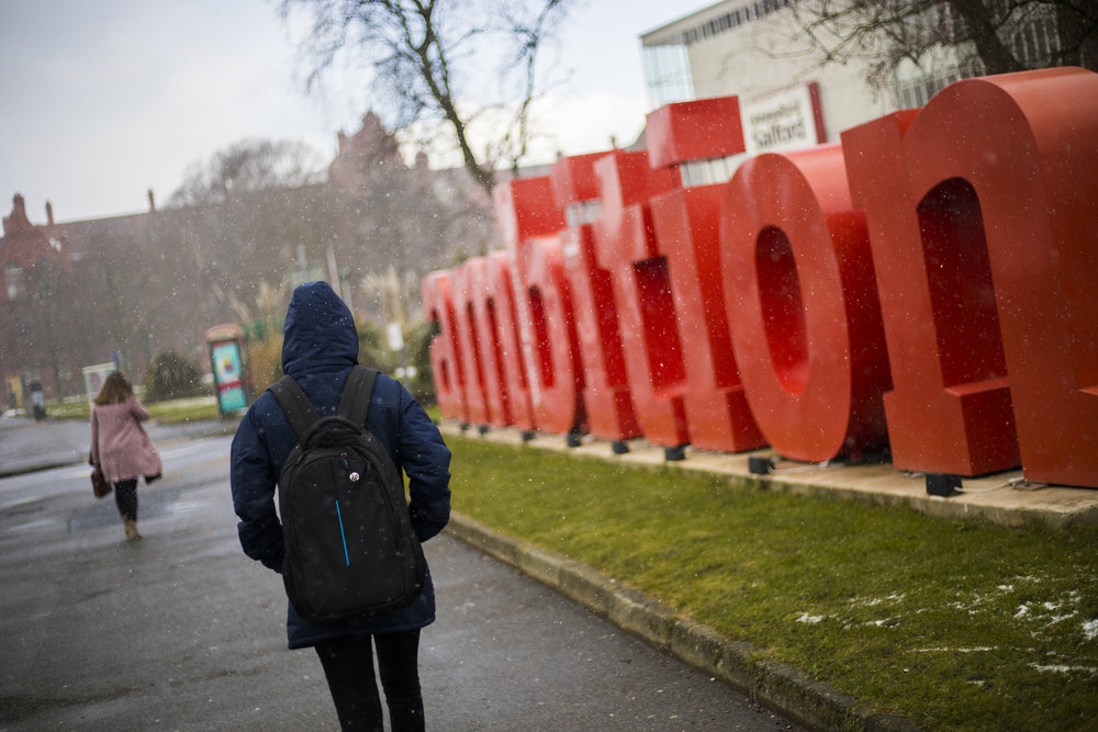 A winter shot of the red 'ambition' sign outside Maxwell building. The photo also shows the back of a student walking past in a dark blue coat. Snow falls around him.