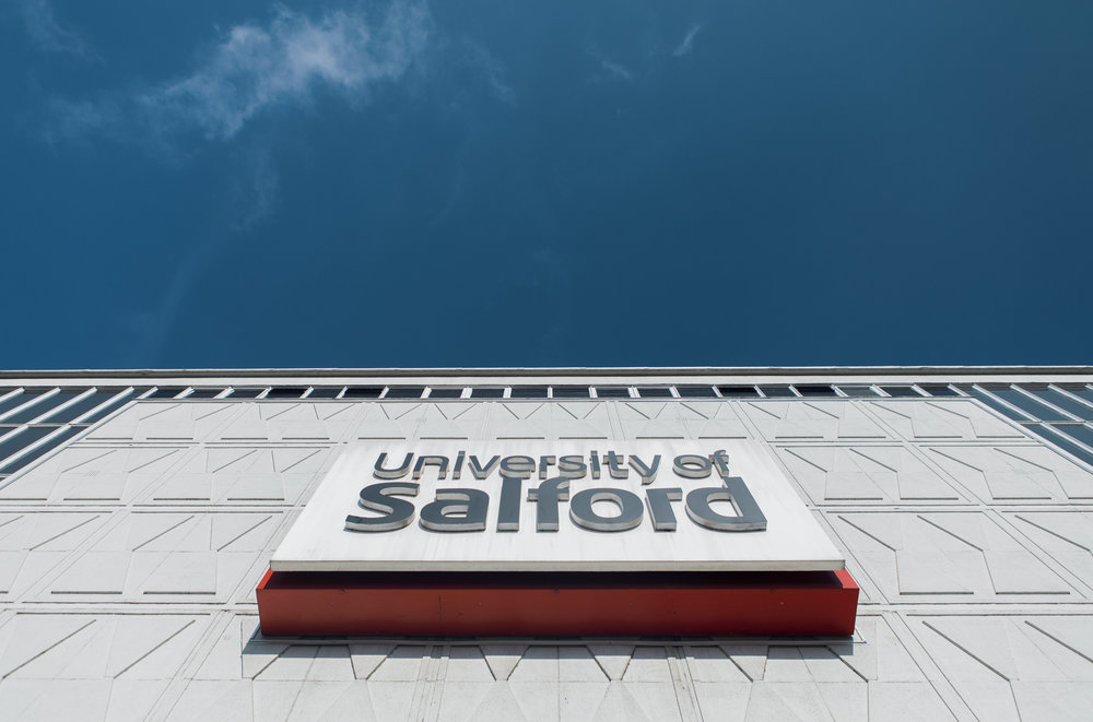 A upward shot of a blue sky and the front of one of the University of Salford buildings that has the logo on it.