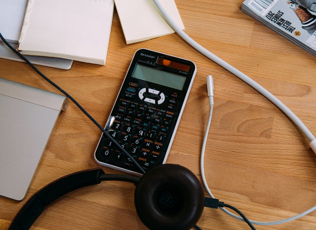 On a desk there is a couple of notebooks, charging wires, a set of headphones and a mathematics calculator.
