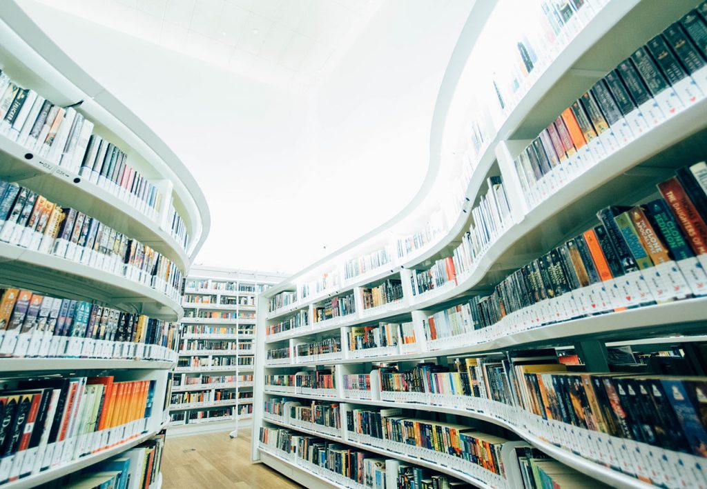 Two winding white framed bookshelves full of books. The floor is a pale laminate wood and in the far background you can see more bookshelves.