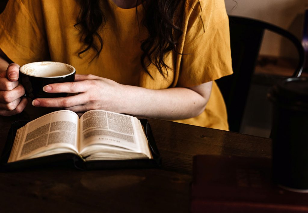 Sat on a dark wooden table is a cup, a book and an opened book that a person is reading. This person wears a mustard yellow t-shirt and has long dark hair. You cannot see their face. They are drinking from a black cup of coffee.