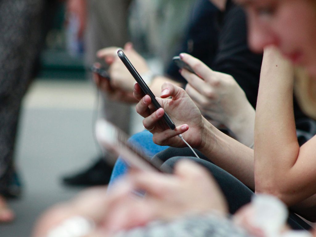 A couple of people sat on a train/tram on their phones. Most of the people in foreground and far background are blurred out aside one person in the middle who is on a black smartphone their hand is in focus, you cannot see their face.