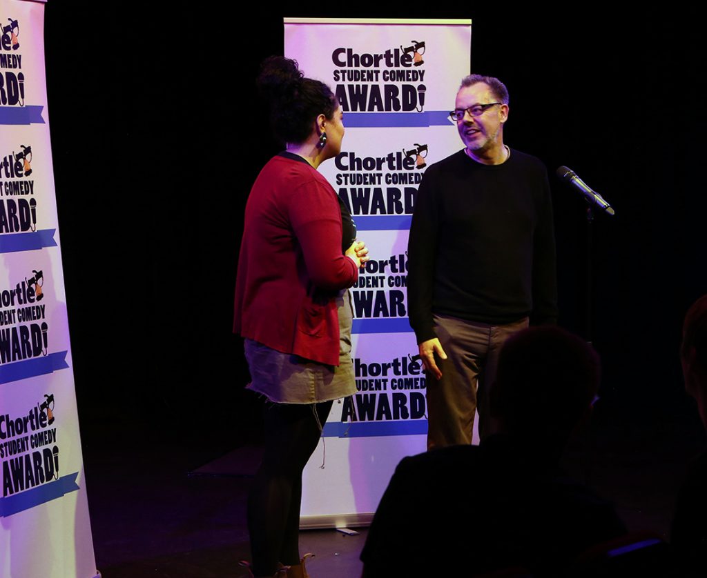 After being announced winner of the Student Comedy Awards, this photo shows Chortle manager Steven Bennett and winner Erika Ehler stood on the stage in front of the microphone talking with the Chortle Student Comedy Award logos behind them