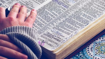 A person in a grey jumper has a detailed book open reading it. The photo also shows that mini pen written notes are dotted across the book. Underneath the open book is a patterned green and blue notebook.