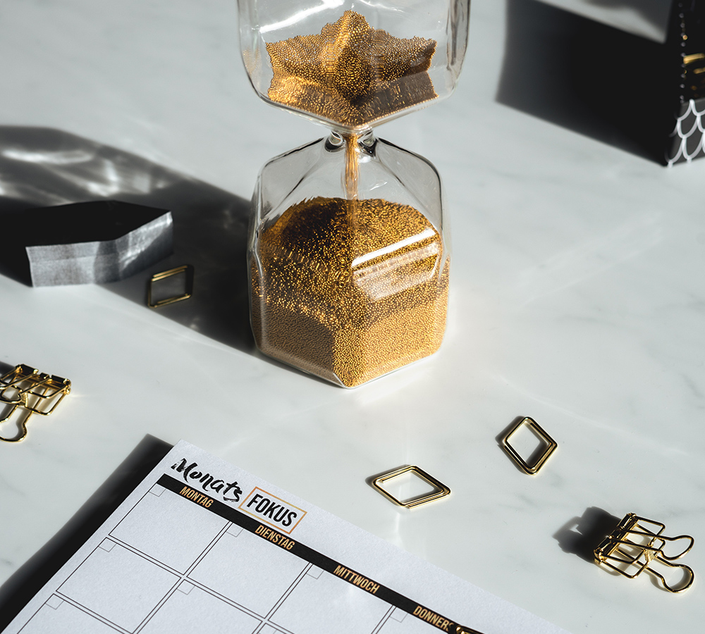 A close up of a white desk that has a sand timer on it with vibrant gold sand. As well as the time there is gold paper clips and binder clips and a desk calendar. 