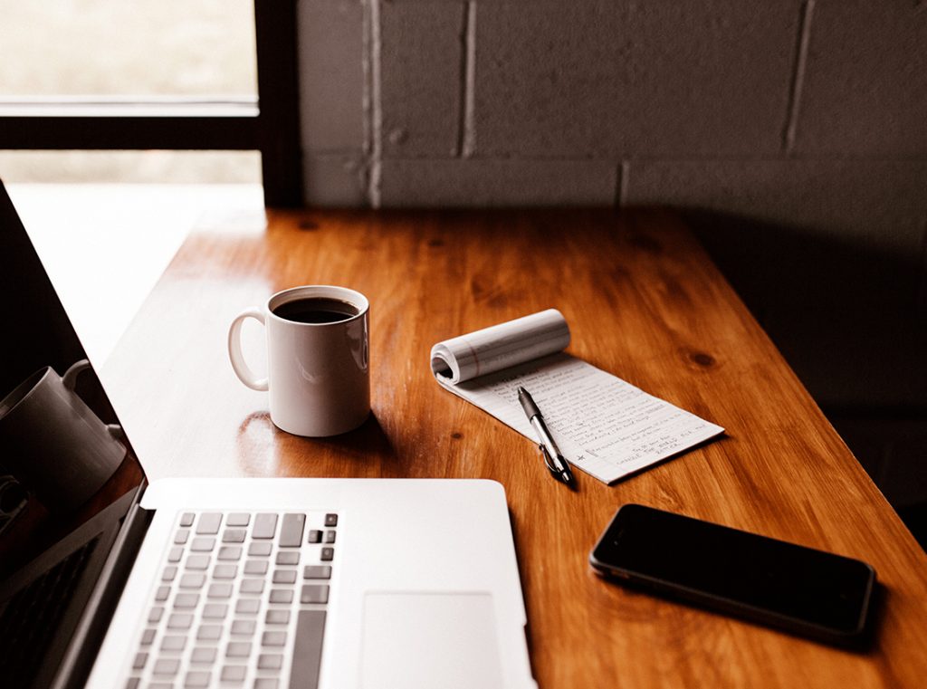 A close up of a wooden desk that has a laptop, smartphone and notepad on it as well as a white mug of black coffee.