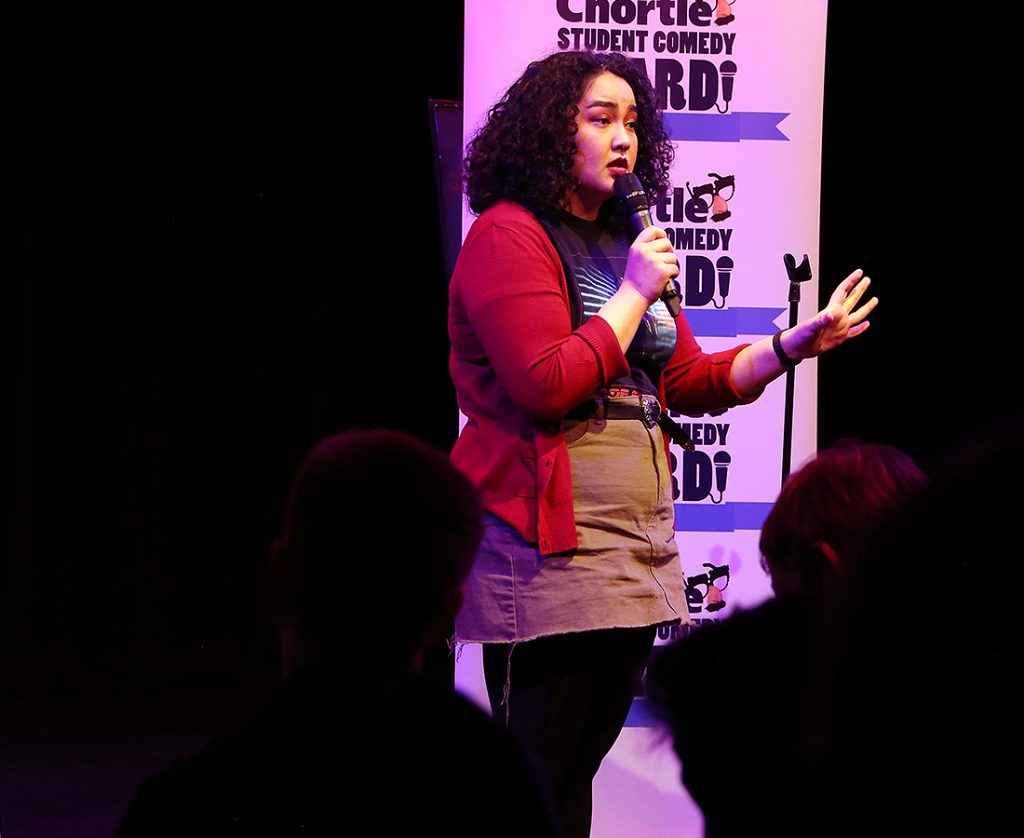 Erika Ehler wearing a red cardigan and khaki skirt, mid-speech performing in front of the crowd. She is holding the microphone and stood in front of one of the flats with the 'Chortle Student Comedy Award' logos on.